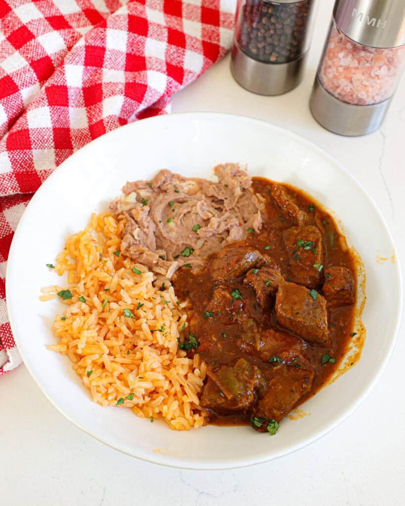 A single serving white bowl with Mexican rice, refried beans, and carne guisada. 