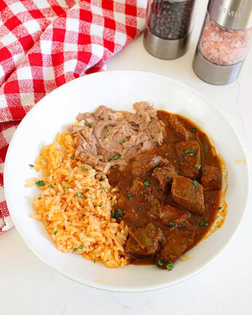 A single serving white bowl with Mexican rice, refried beans, and carne guisada.