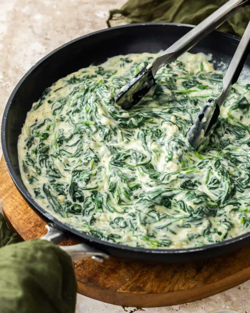 A skillet full of creamed spinach with a pair of tongs in the skillet.
