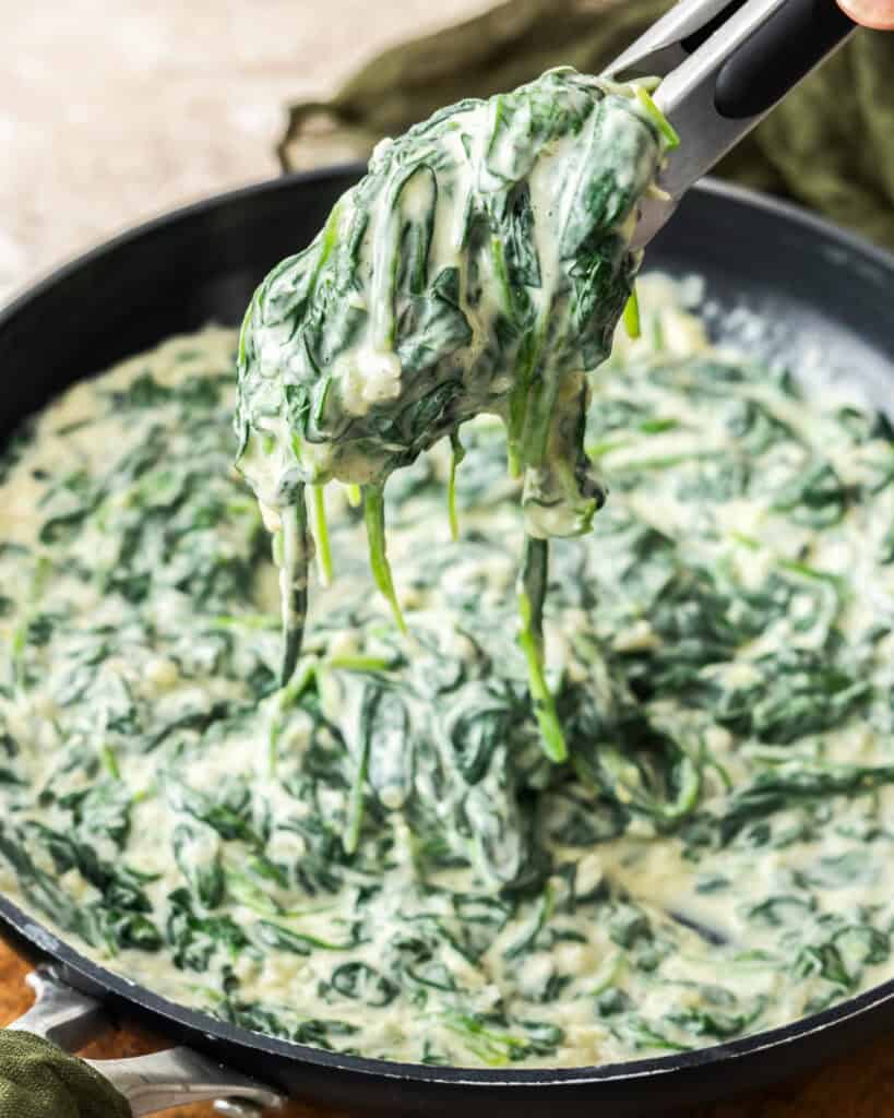 Tongs full of homemade creamed spinach over a large skillet.