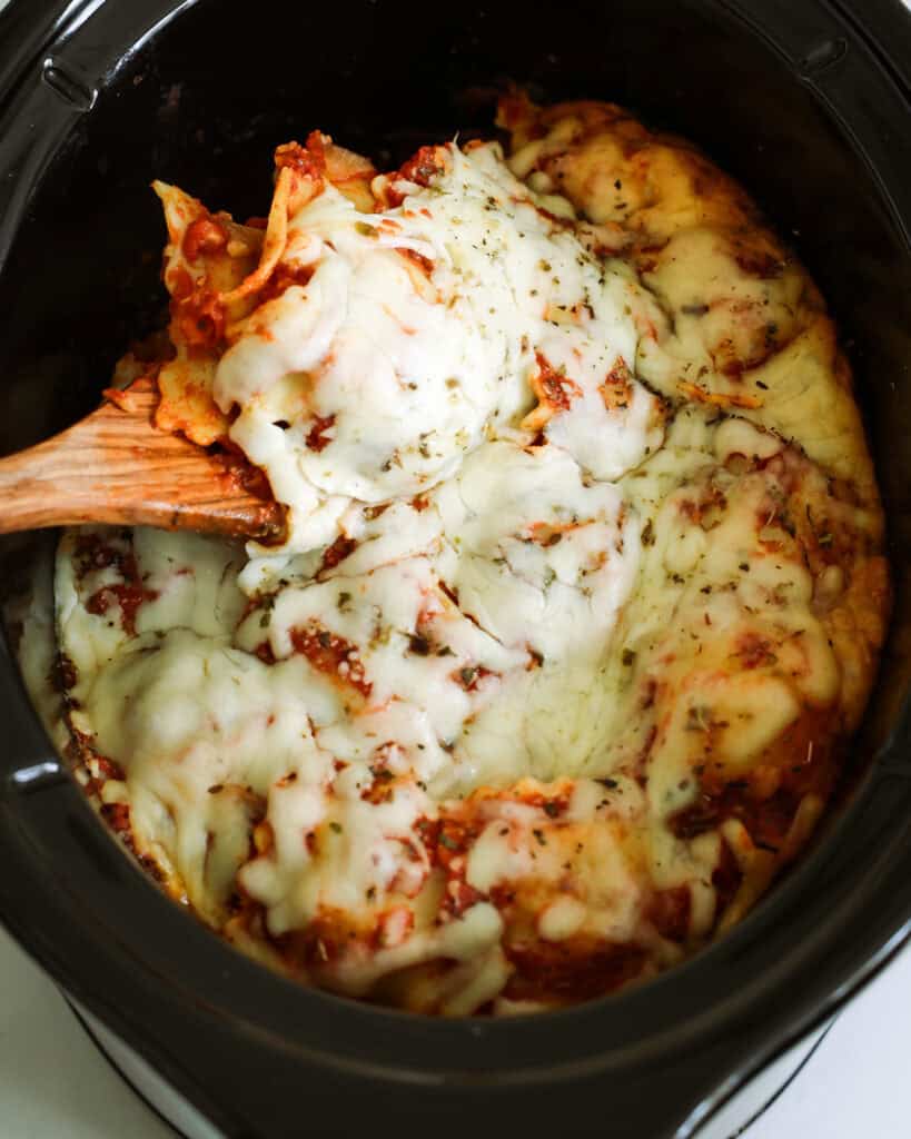 An overhead view of a crock pot and wooden spoon full of ravioli lasagna. 