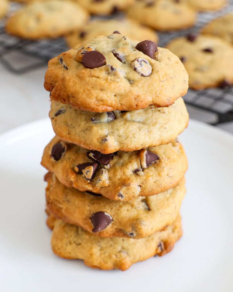 A stack of banana chocolate chip cookies on a plate in front of a wire cooling rack full of cookies. 
