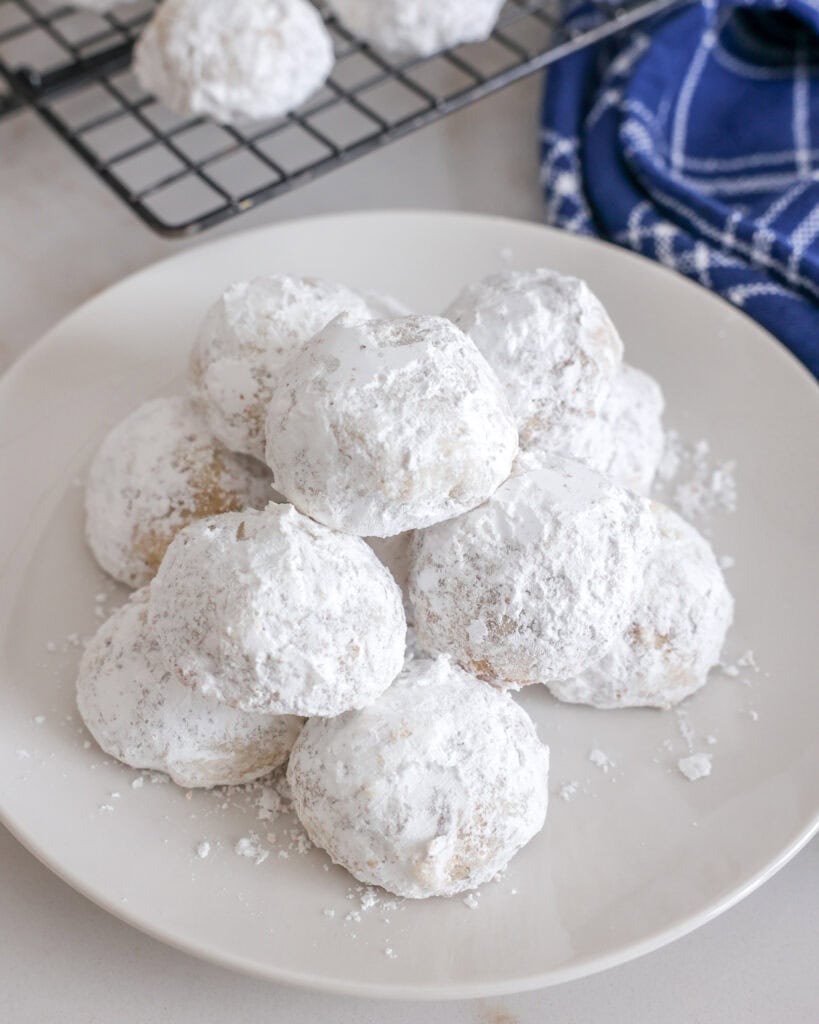 A white plate full of Mexican wedding cookies with a blue towel and a cooling rack in the background. 