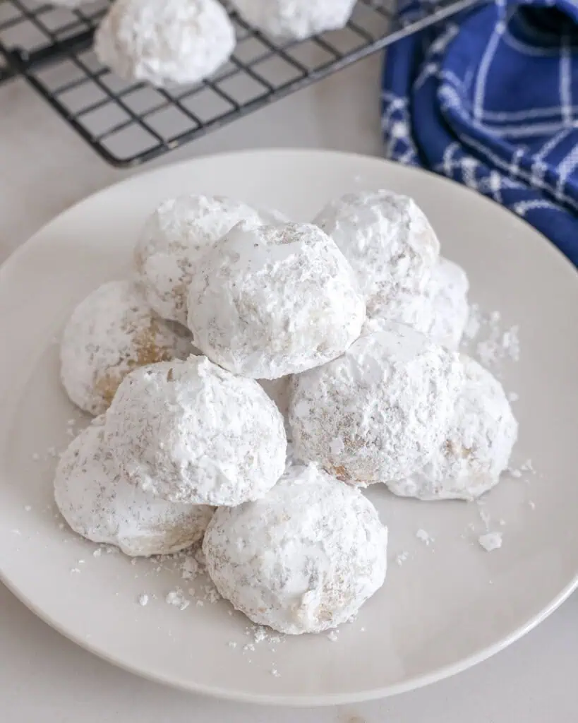 A white plate full of Mexican wedding cookies with a blue towel and a cooling rack in the background.