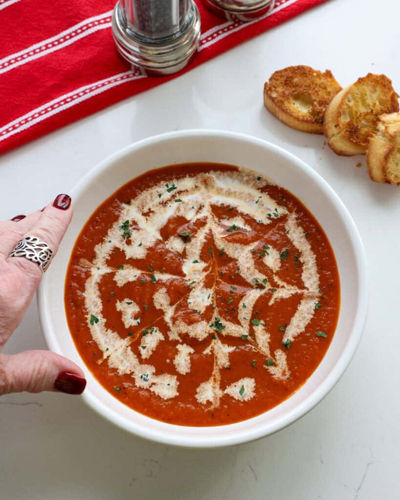 A single serving bowl full of roasted red pepper soup garnished with cream and chopped fresh parsley. 