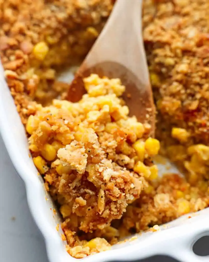 A close up view of scalloped corn with a buttery cracker topping on a wooden spoon.