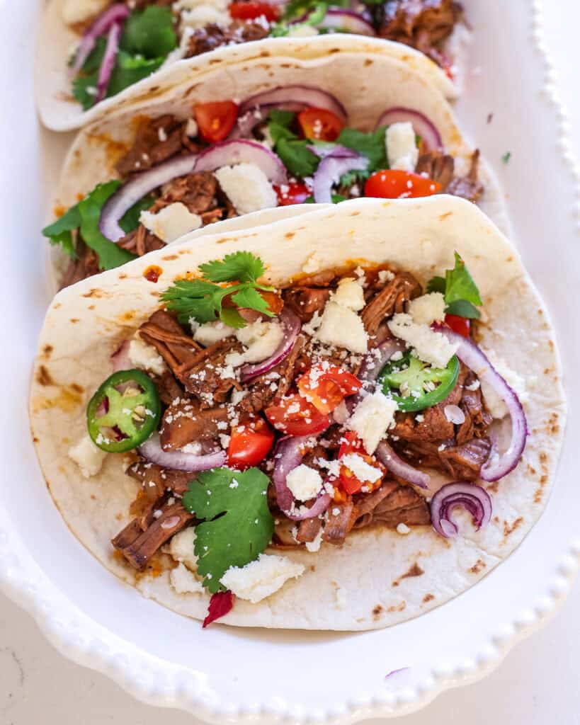 A close up view of shredded beef tacos with jalapenos, tomatoes, red onion, cilantro, and Cotija cheese. 