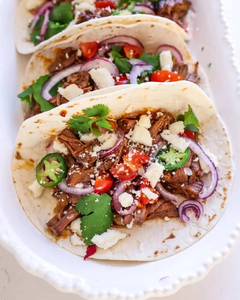 A close up view of shredded beef tacos with jalapenos, tomatoes, red onion, cilantro, and Cotija cheese.
