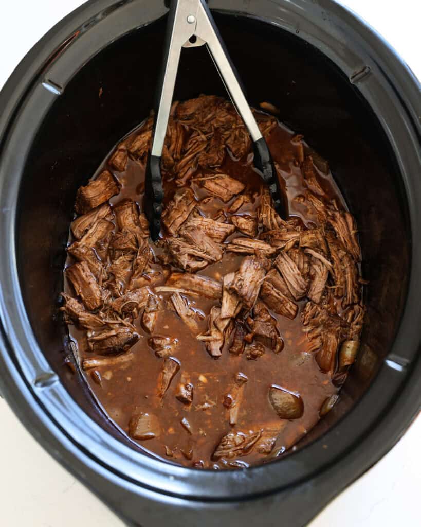 Shredded beef taco meat in a crock pot with a pair of tongs. 