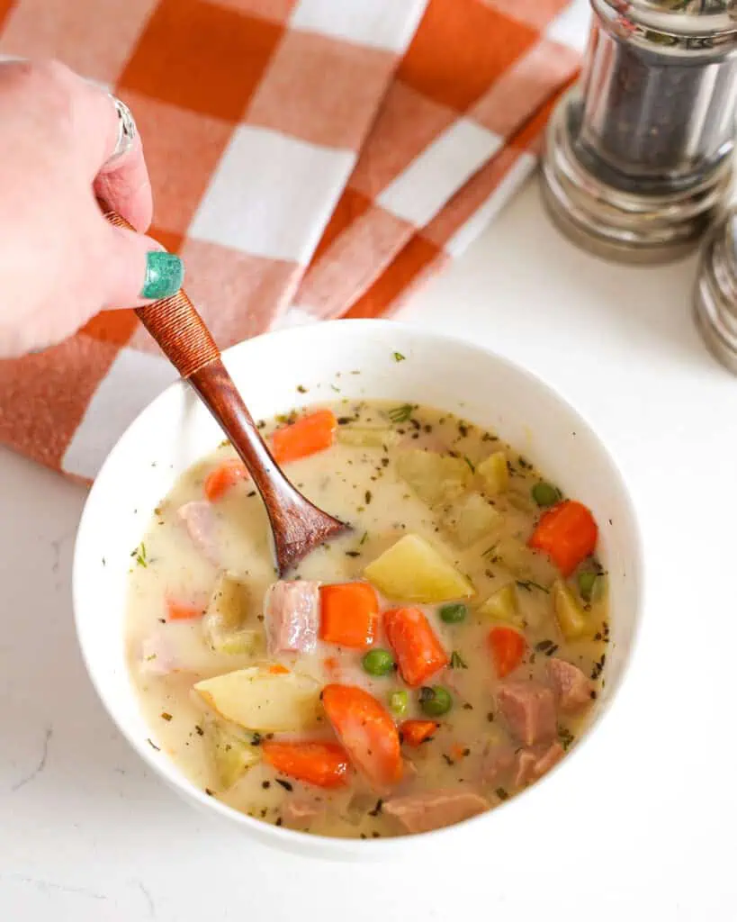A single serving bowl with a wooden spoon in it full of crockpot ham and potato soup with a salt and pepper shaker in the background.