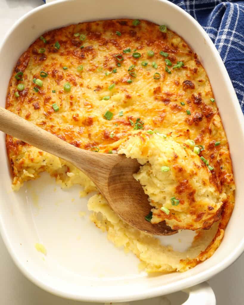 An overhead view of an oval casserole dish and a wooden spoon full of cheesy mashed potatoes. 