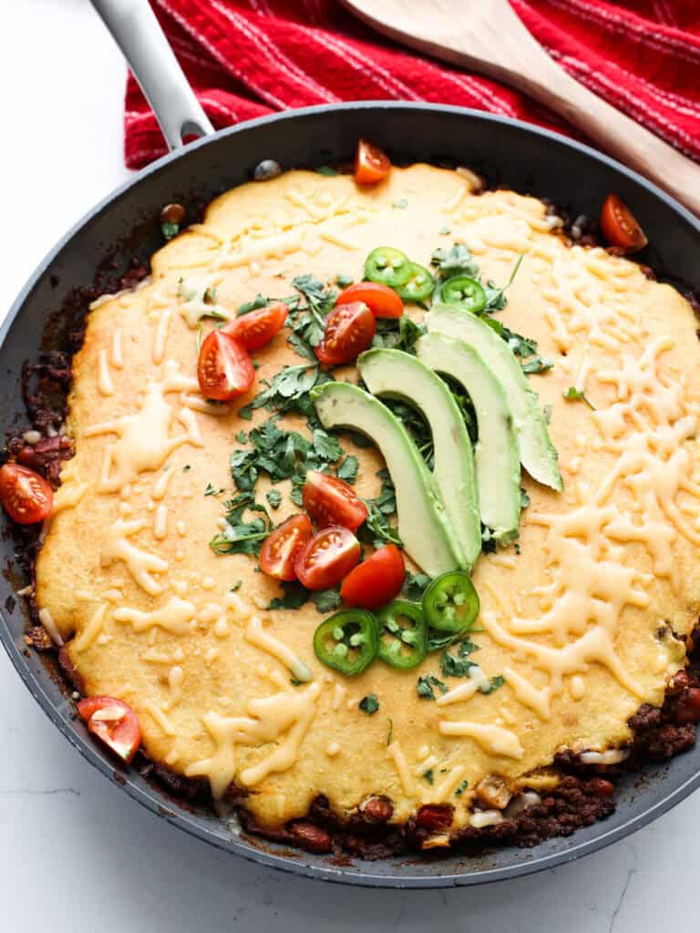 A large skillet full of chili cornbread casserole topped with jalapenos, tomatoes, cilantro, and avocado. 