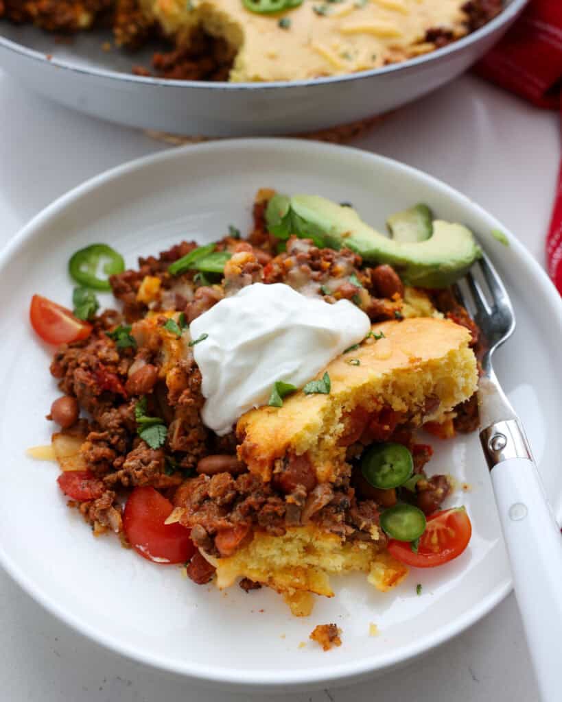 A single serving plate full of chili cornbread casserole topping with tomatoes, jalapeno, avovcado, and sour cream. 
