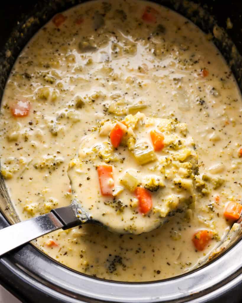 A crockpot and a large ladle full of crockpot broccoli cheddar soup. 