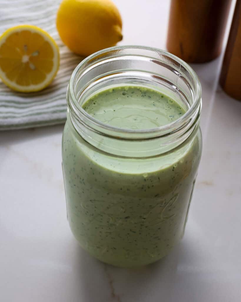 A mason jar full of Green Goddess dressing with lemons and a salt and pepper shaker in the background. 
