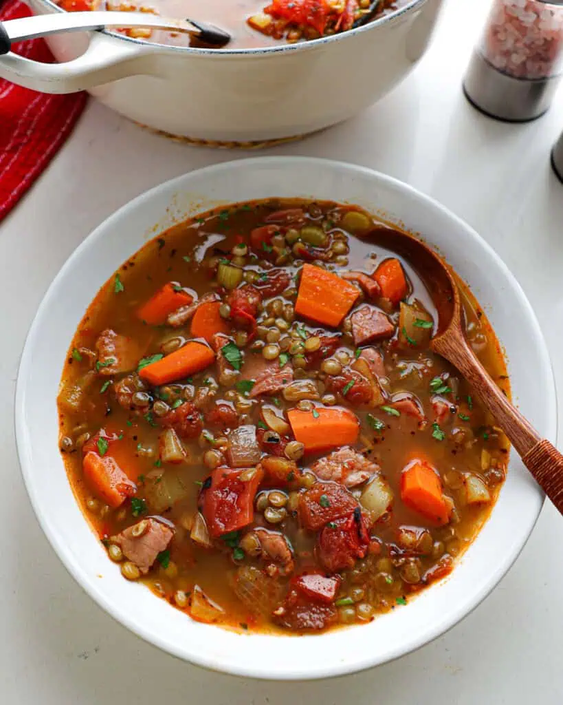 A serving bowl with a wooden spoon in it full of ham and lentil soup.