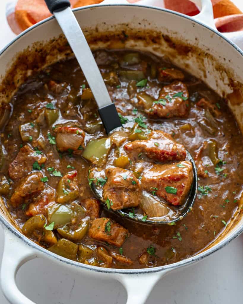 A close up view of a ladle full of beef tips over a Dutch oven full of beef tips. 