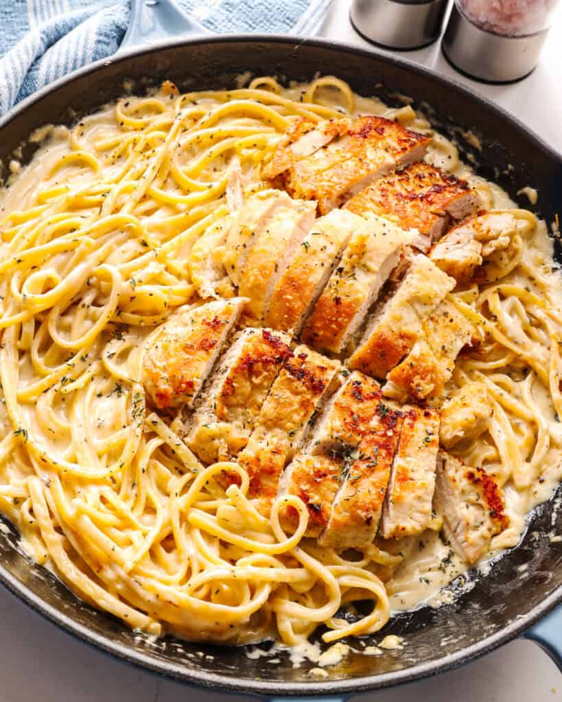 A cast iron skillet full of creamy alfredo pasta with sliced golden brown chicken on top. 