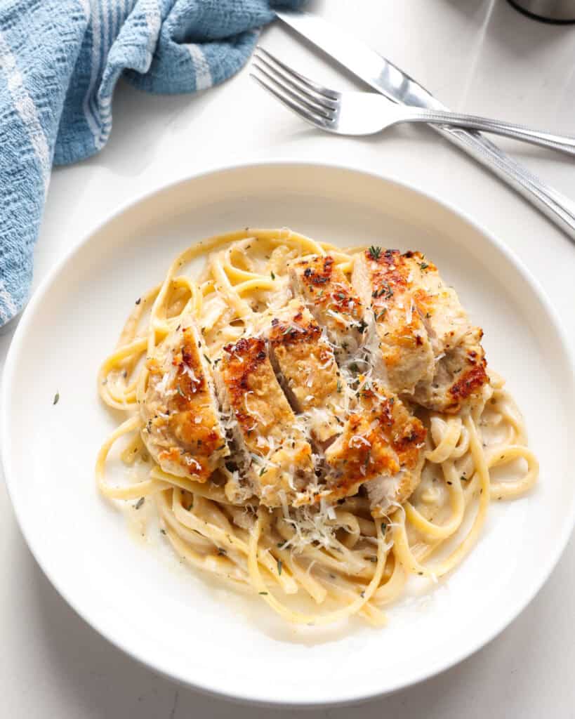 A single serving white plate full of creamy chicken pasta with a fork and knife in the background with a blue kirchen towel. 