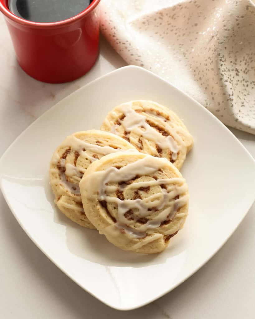 Three cinnamon roll cookies on a single serving white plate with a coffee cup in the background. 