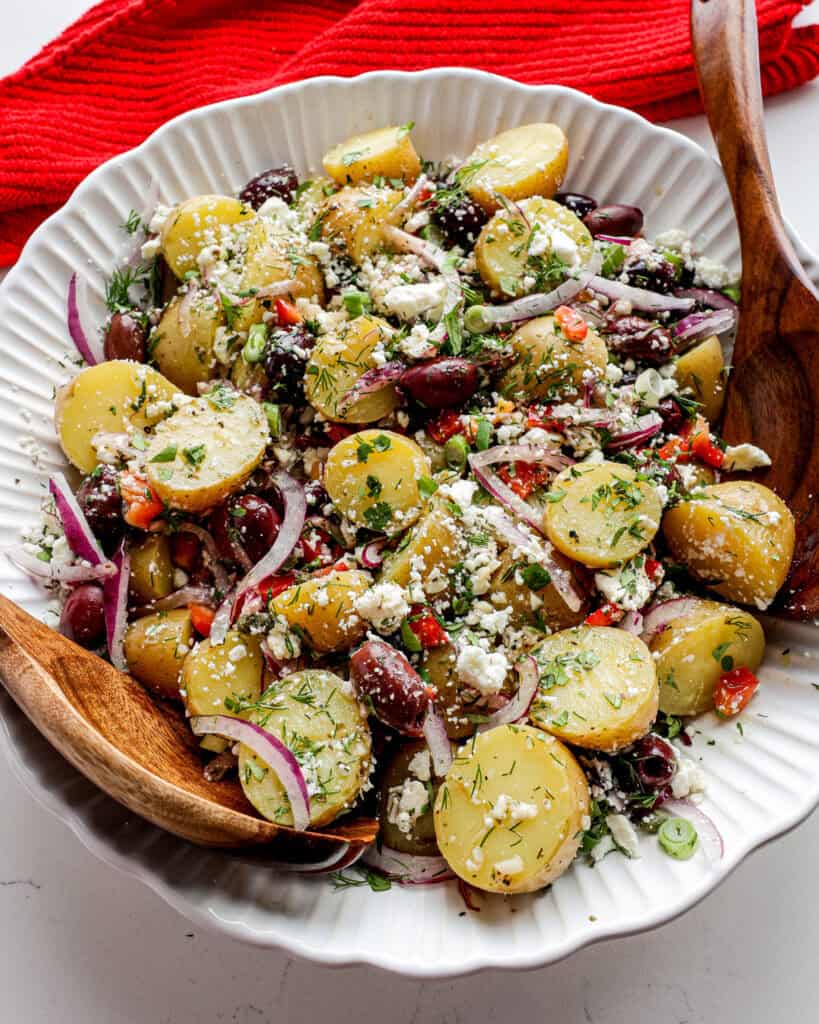 A large white serving bowl full of Greek potato salad with wooden serving spoons. 