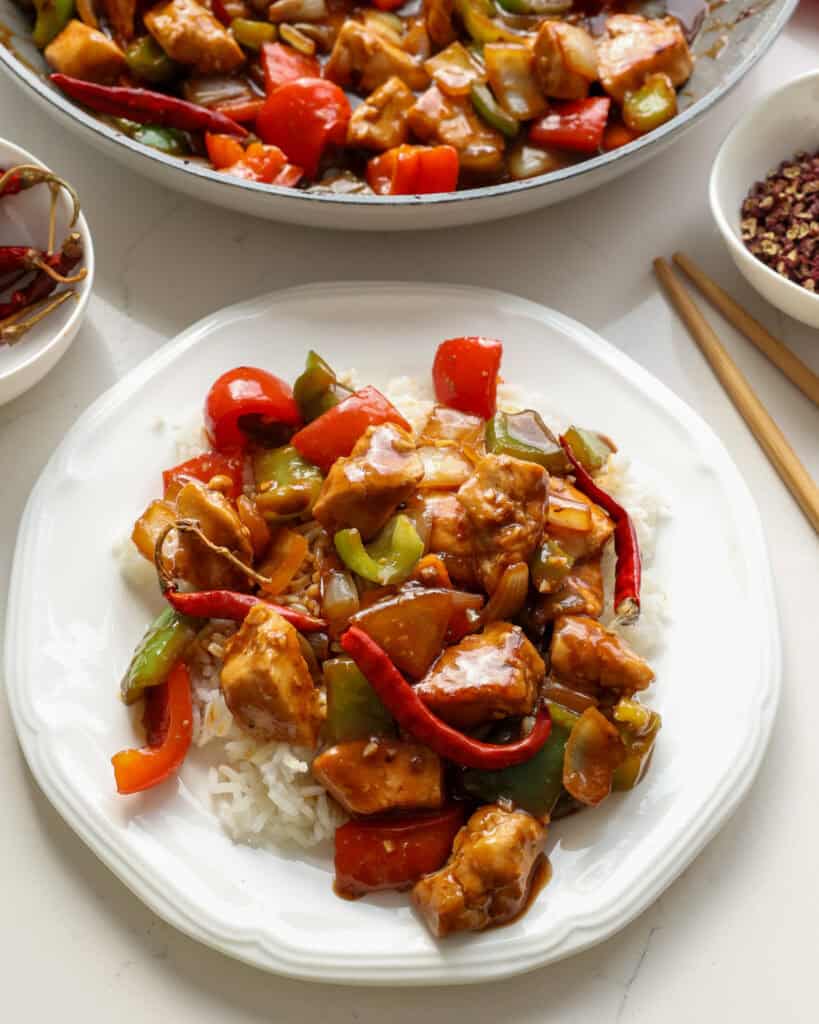 A plate full of Szechuan chicken over rice with a skillet full of szechuan chicken in the background. 