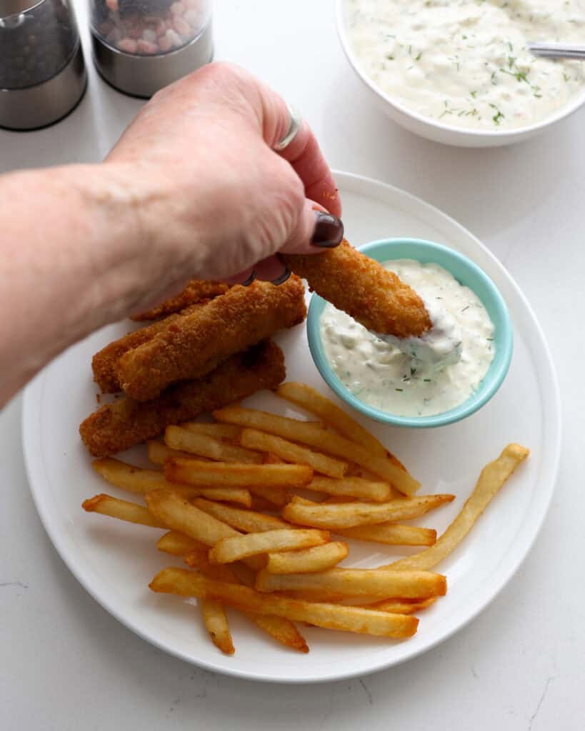 A fish stick dipped in tartar sauce on a plate with fish sticks and French fries.