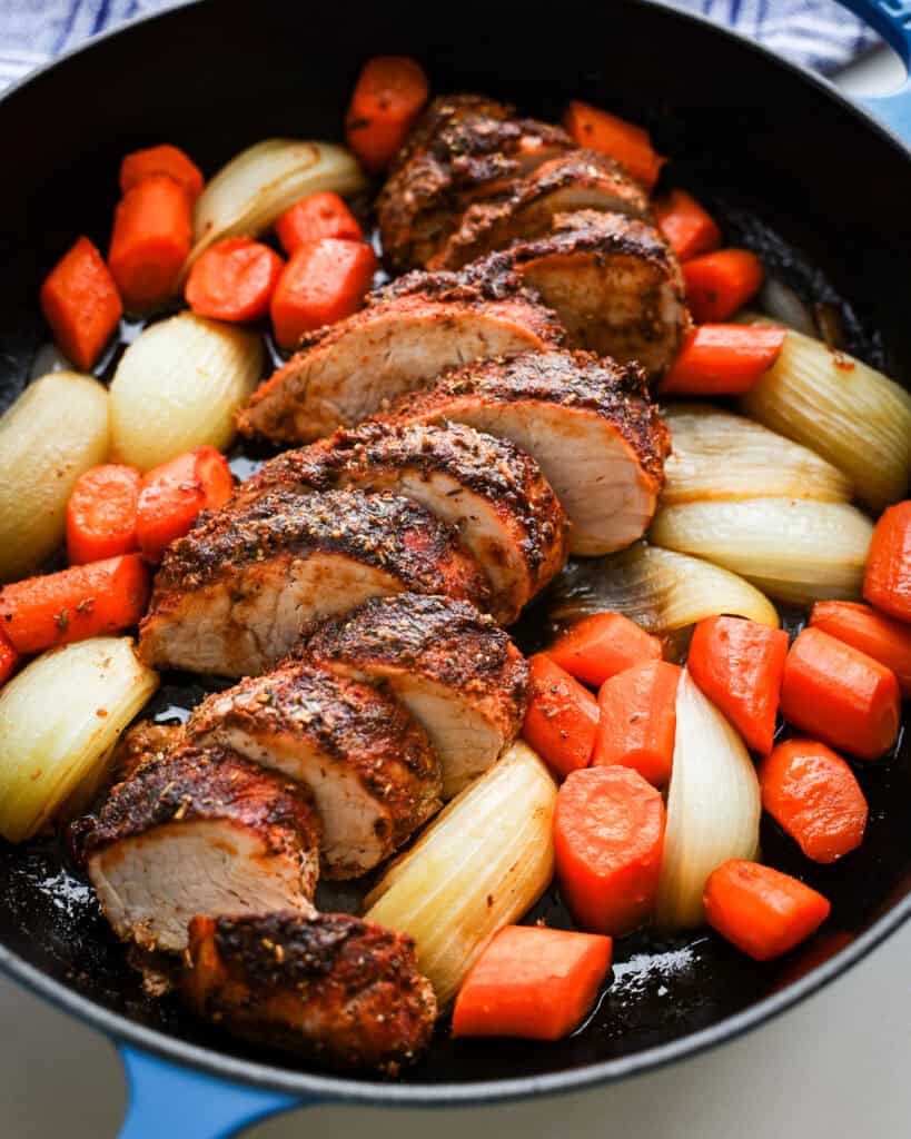 A close up view of pork tenderloin with onions and carrots in a cast iron skillet. 