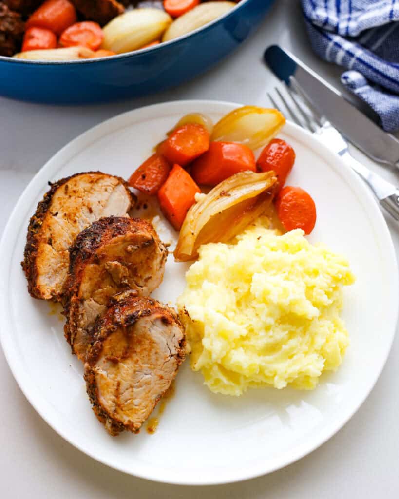 A single serving white plate with baked pork tenderloin, carrots, onions, and mashed potatoes. 