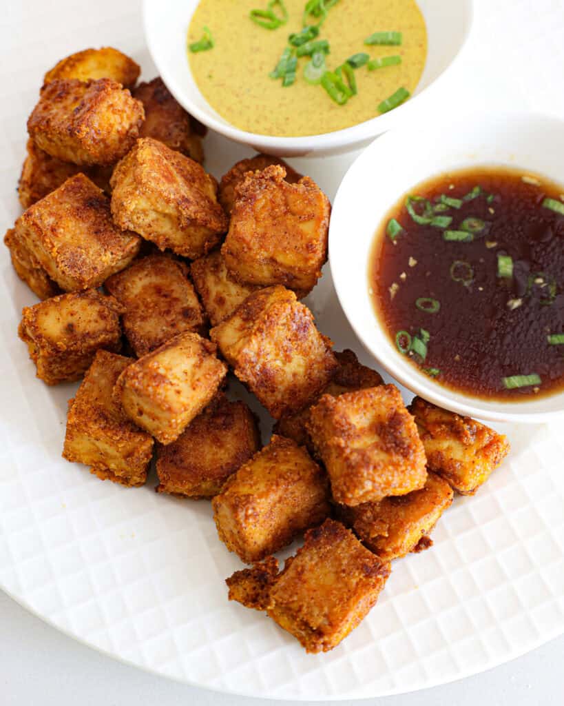 A close up view of crispy fried tofu with a a bowl of spicy mustard and a bowl of ginger soy sauce. 
