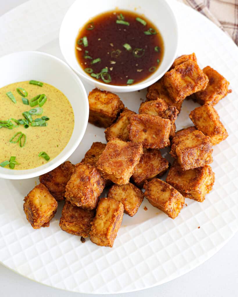 An overhead view of crispy tofu on a white serving plate with bowls of spicy mustard and ginger soy sauce. 