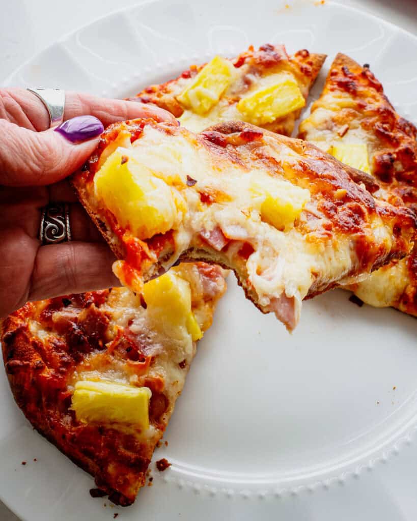 An individual piece of Hawaiian pizza with a bite out of it. 