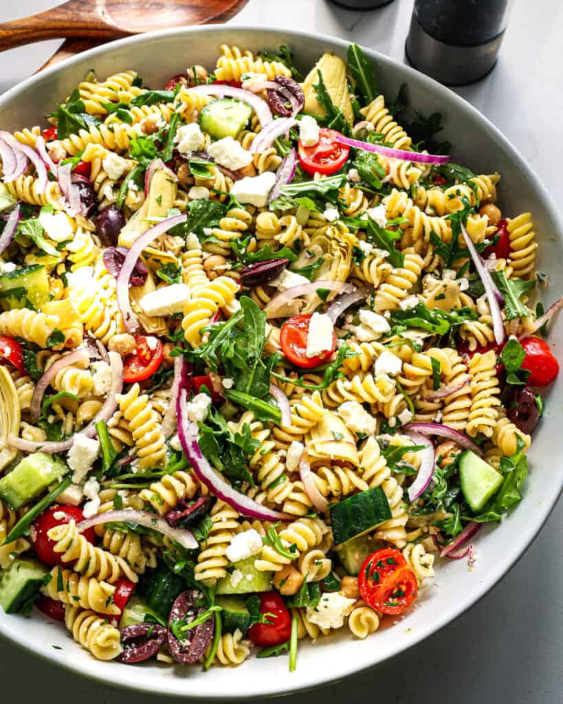 A close up view of a large white serving bowl full of Mediterranean pasta salad. 