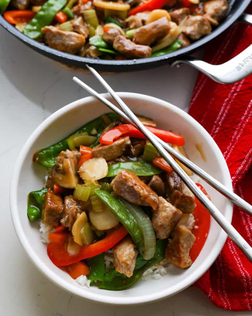 A single serving bowl full of pork chop suey over rice with a pair of chopsticks on the bowl and the skillet full of chop suey in the background. 