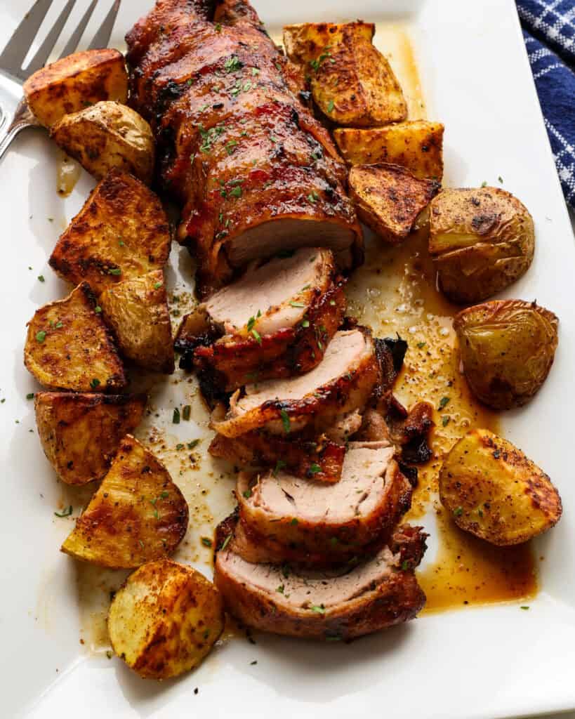 A large white serving platter with bacon wrapped porked tenderloin and over roasted potatoes. 