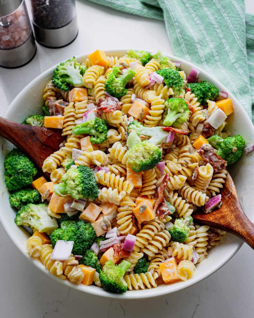 An overhead view of broccoli pasta salad in a large white bowl with wooden serving pieces.