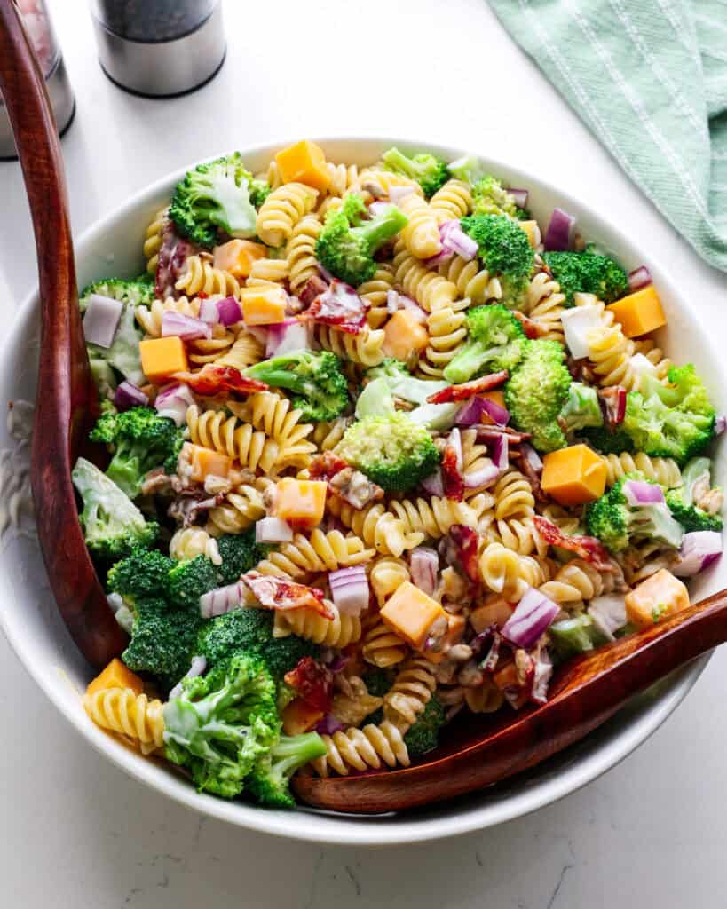 A close up view of broccoli pasta salad in a whitle bowl with wooden serving tongs.