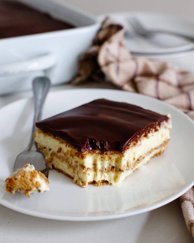 A slice of chocolate eclair cake on a single serving white plate with a fork with the rest of the cake in the background. 