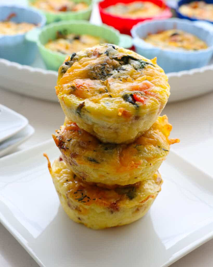 Three egg bites stacked on a white serving plate with more eggs bites in the background. 