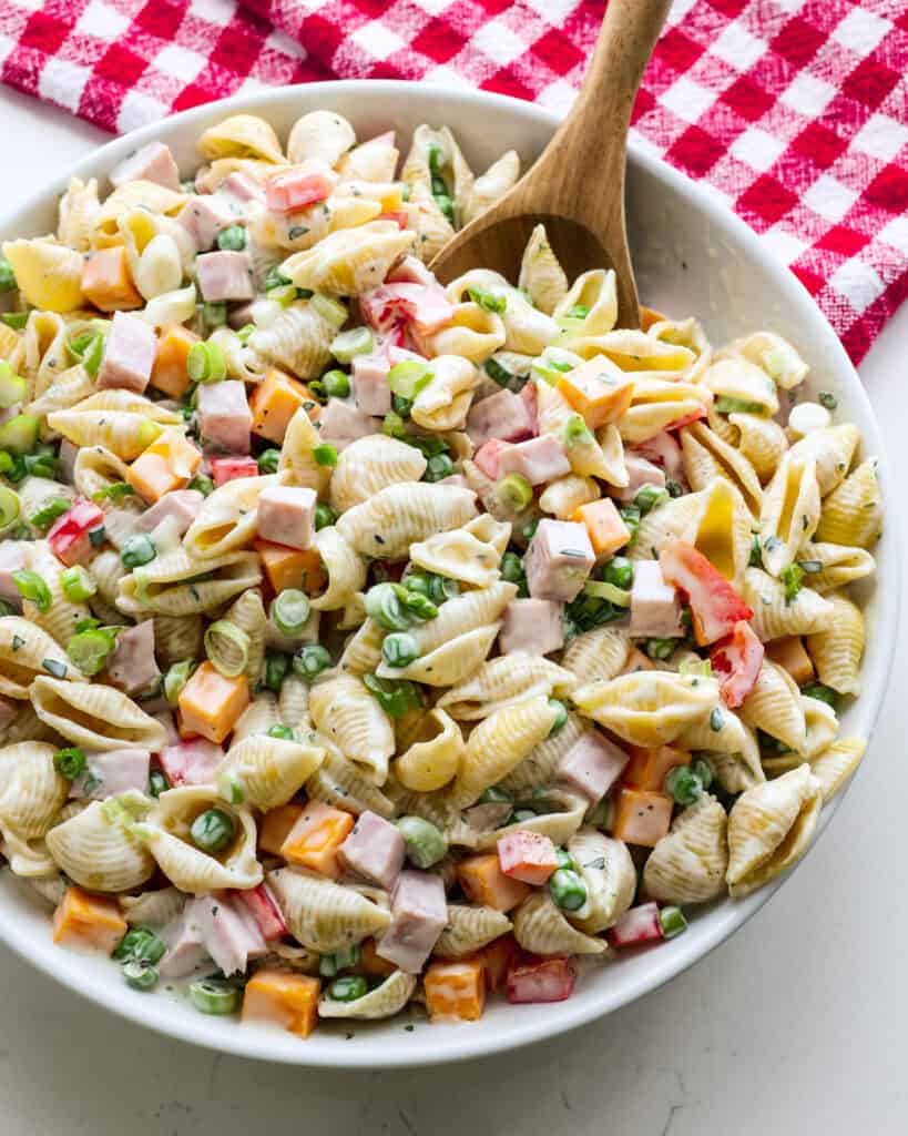 A serving bowl and a wooden spoon full of ham pasta salad. 