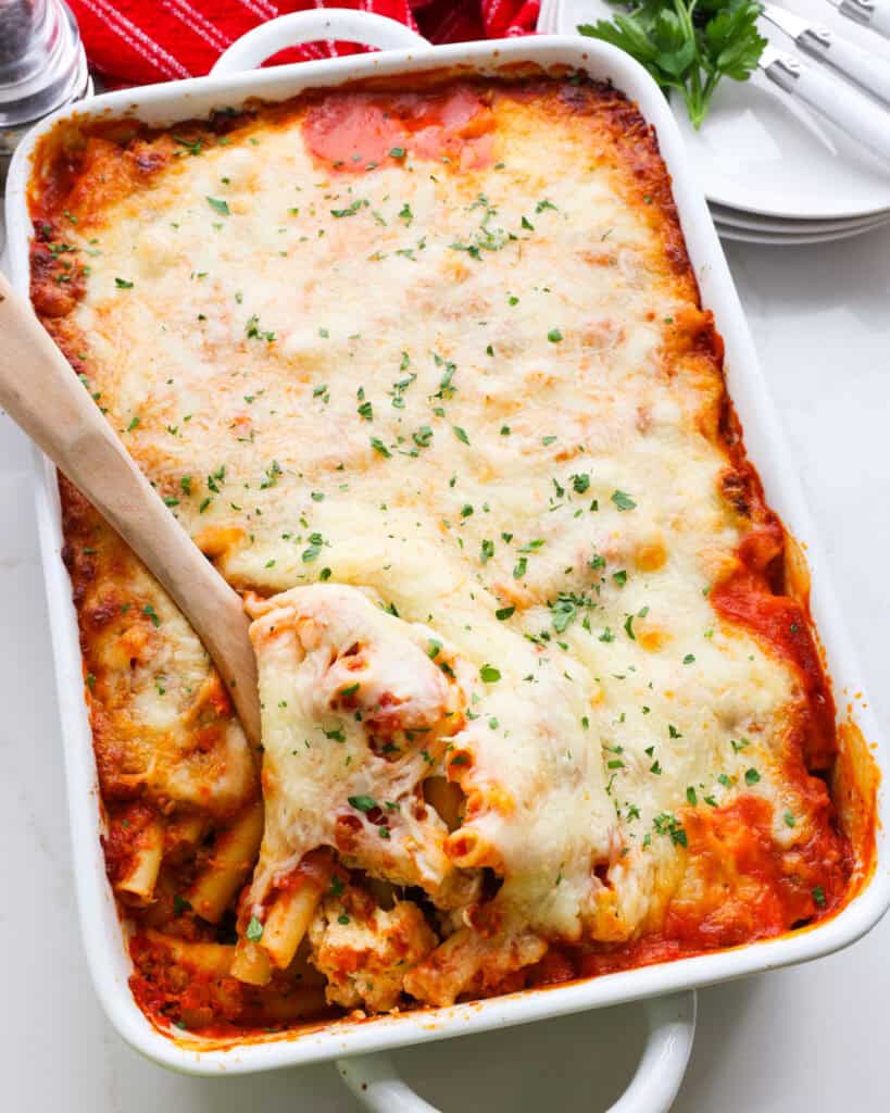 Baked Ziti in a white casserole dish with a wooden spoon. Plates and forks are in the background. 