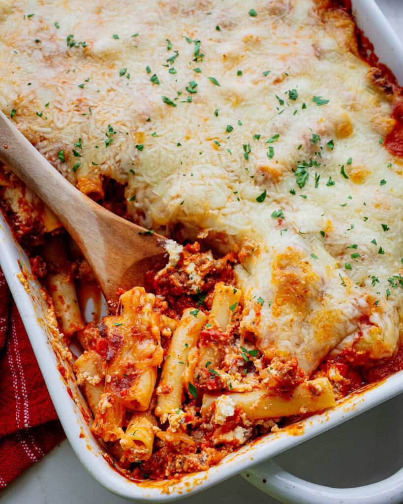 A close up view of baked ziti on a wooden spoon in a white casserole dish. 
