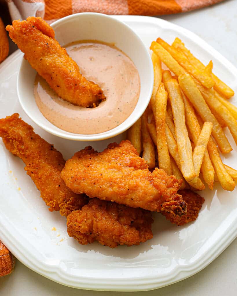 A chicken tender in a bowl full of copycat cane's sauce on a plate full of chicken tender and French fries. 