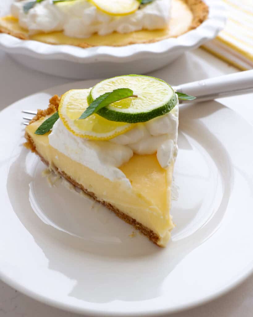 A slice of lemon cream pie on a white serving plate with a fork. The rest of the pie is the the background.