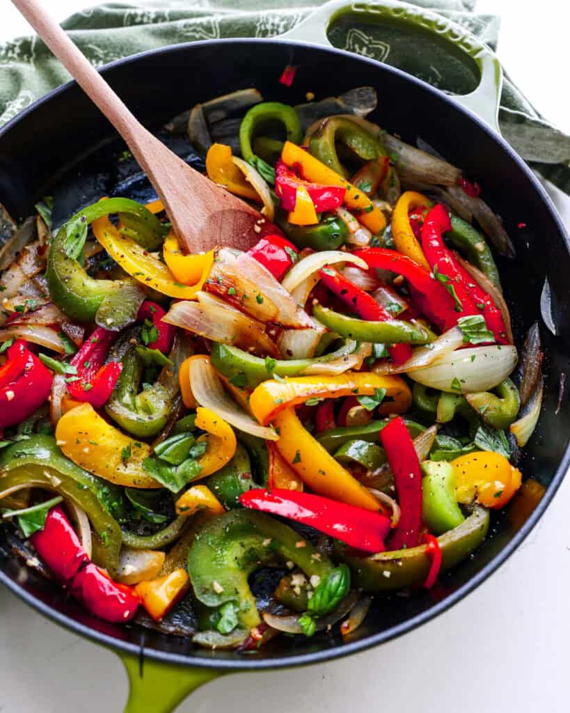 An overhead view of sauteed peppers and onions in a skillet with a wooden spoon.