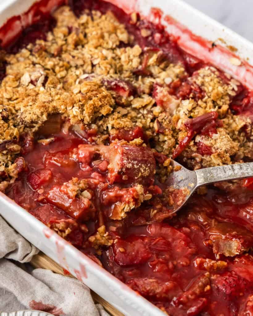Strawberry Rhubarb Crumble Recipe Overhead shot of Strawberry Rhubarb Crisp with a spoon in the white dish and strawberries to the left.