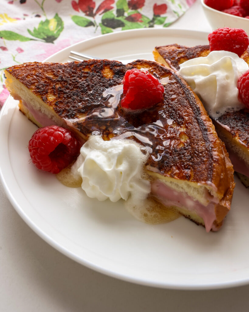 A close up view of a piece of stuffed French toast with raspberries, whipped cream, and maple syrup. 