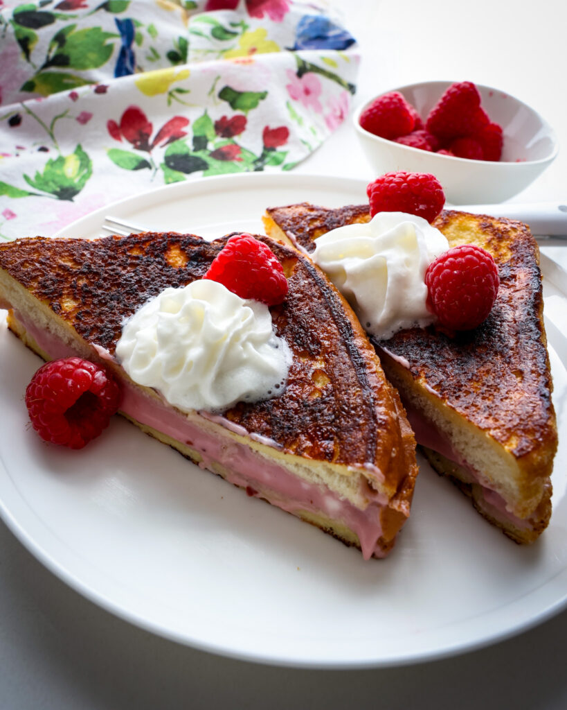 Two pieces of stuffed French Toast topped with raspberies and whipped cream on a white serving plate. 