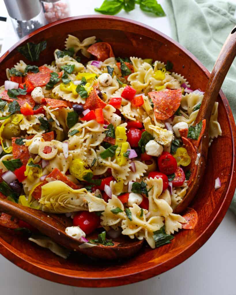 A close up view of antipasto pasta saald in a wooden bowl with wooden untensils.