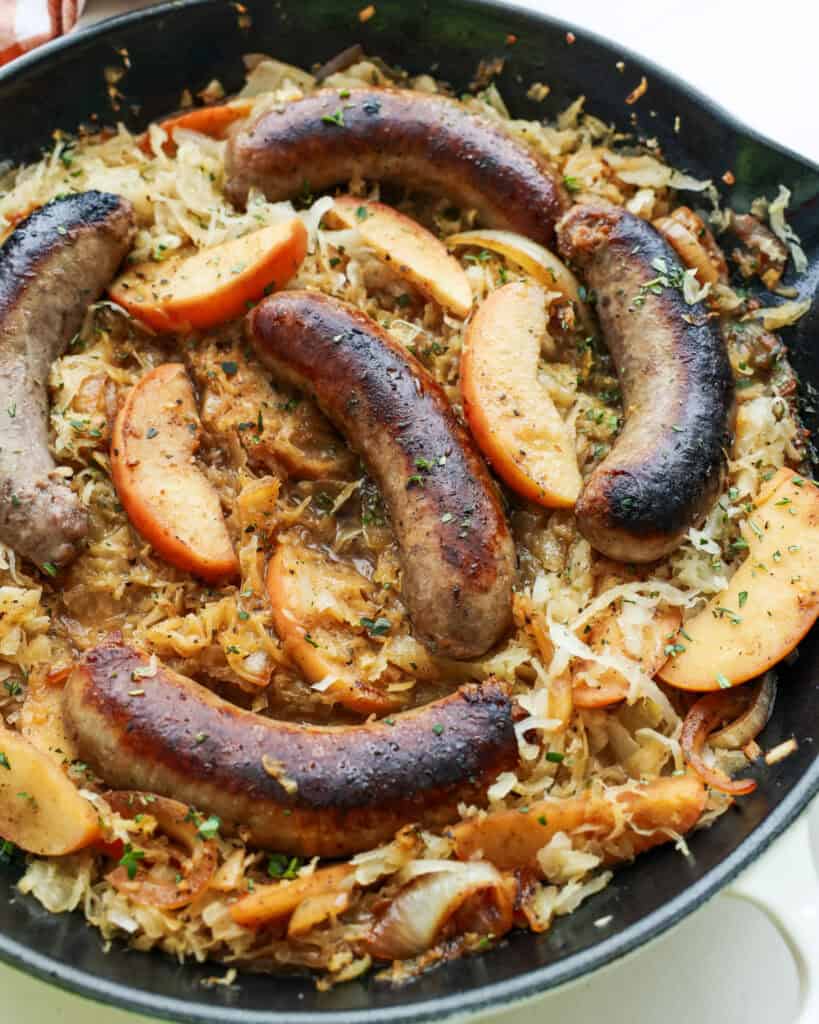 An overhead view of bratwurst, sauerkraut, and apples in a white skillet. 
